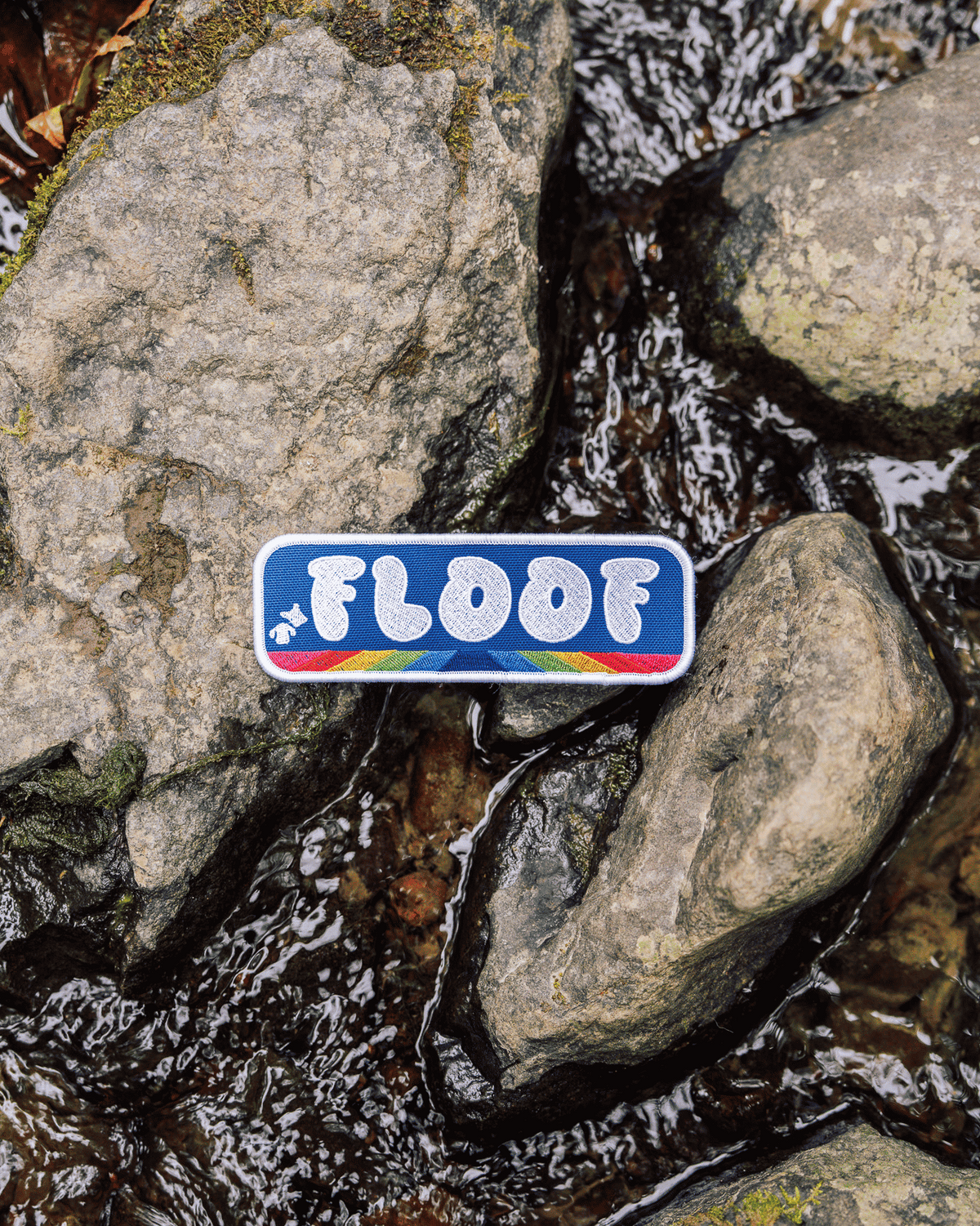 A rectangular enamel pin with the word “FLOOF” in white letters on a blue background, outlined in white and accented with small red, yellow, and blue stripes, placed on wet rocks near a stream.