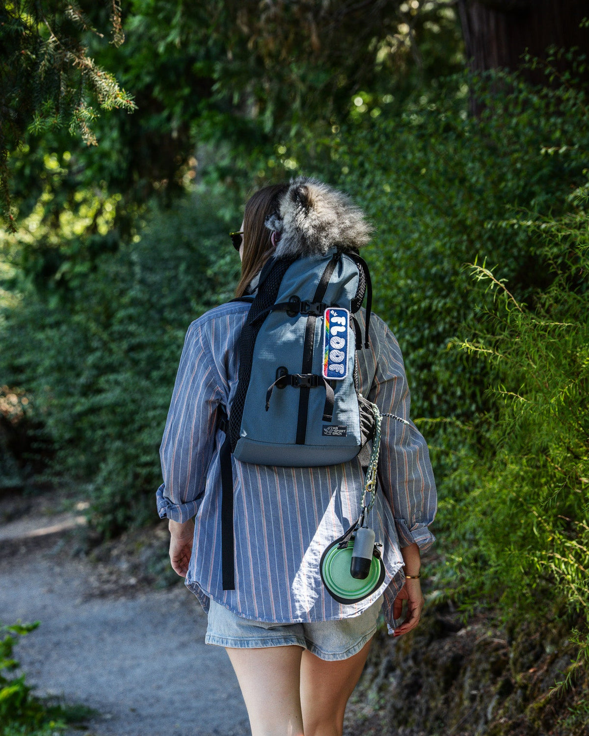 A woman seen from behind walking on a forest path, wearing a gray backpack with a fur-trimmed hood, a blue-and-white striped shirt, and denim shorts.