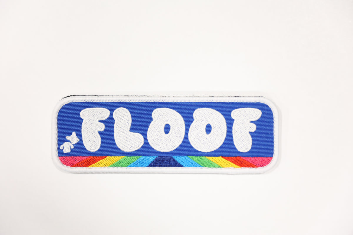 An embroidered rectangular patch with rounded corners featuring the word 'FLOOF' in white bubble letters on a blue background, with a small white dog silhouette and a rainbow stripe detail along the bottom edge.